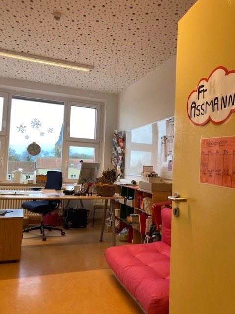 Im vorderen Bereich des Büros steht ein rotes Sofa, weiter hinten ein Schreibtisch, ein Bücherregal und ein Whiteboard an der Wand.